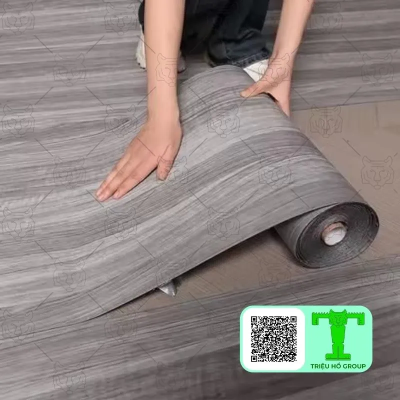 Người đang trải cuộn sàn nhựa vinyl vân gỗ màu xám lên sàn nhà, cho thấy quá trình thi công dễ dàng