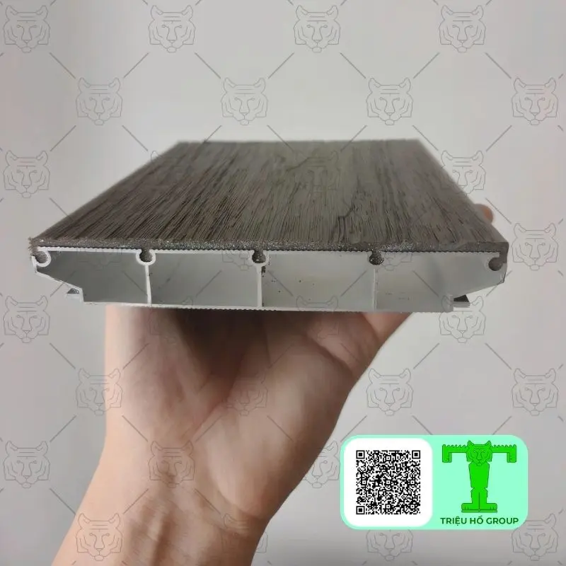 Mặt cắt ngang của tấm sàn nhựa vân gỗ cho thấy cấu tạo nhiều lớp và các khoang rỗng bên trong, được một bàn tay giữ