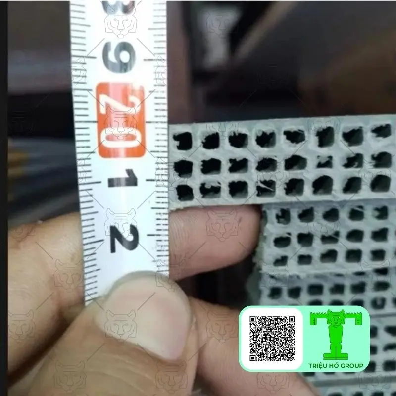 Một bàn tay giữ thước đo bên cạnh tấm ván nhựa cốp pha lõi rỗng màu xám nhạt, cho thấy cấu trúc bên trong và độ dày của vật