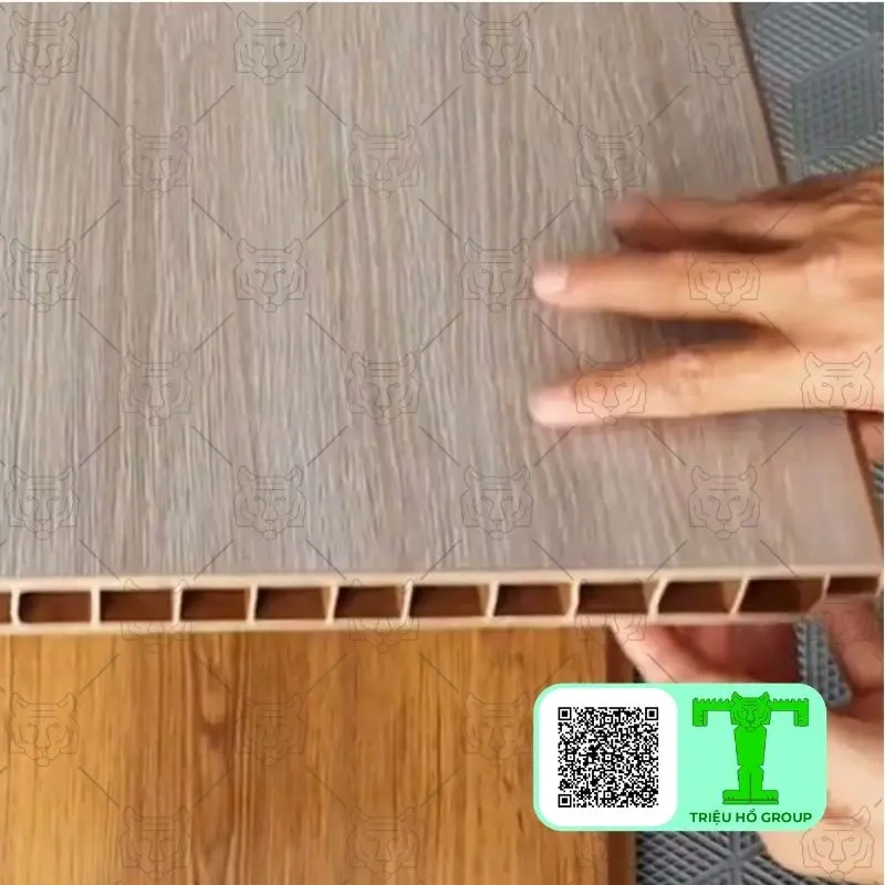 Bàn tay người giữ tấm ván sàn nhựa PVC rỗng ruột vân gỗ màu xám nhạt, cho thấy cấu trúc bên trong